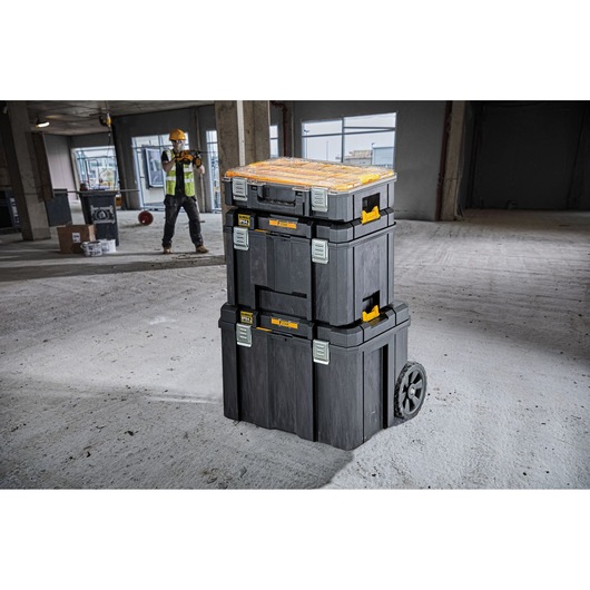 Stacked DEWALT tool boxes with wheels in a construction area, worker in background wearing safety gear.