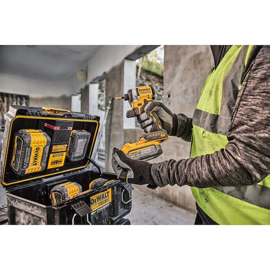 A person wearing gloves and a high-visibility vest is holding a DEWALT cordless power tool with a battery attached. Nearby
