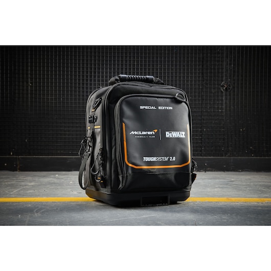 Black DEWALT ToughSystem 2.0 McLaren edition backpack placed on a concrete floor near a black wire wall.