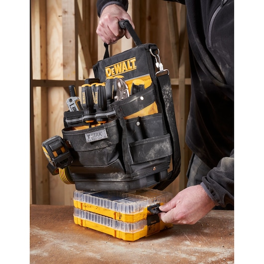 A DEWALT tool bag filled with various hand tools is being carried by a person. The person is placing the tool bag onto two