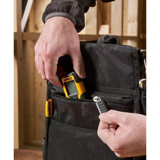 A close-up of hands placing a DEWALT digital tool and a small accessory into the front pocket of a black DEWALT tool bag