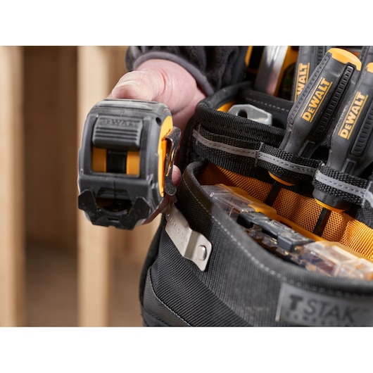 Close-up of a DEWALT tool belt with a person holding a DEWALT tape measure. Several DEWALT screwdrivers and other tools are