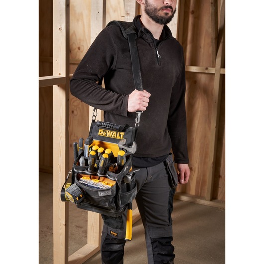 A person wearing work clothes holds a DEWALT tool bag filled with various hand tools and accessories. The tool bag is black