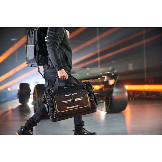 Person holding DEWALT ToughSystem 2.0 tool bag with McLaren branding, walking near a race car indoors.