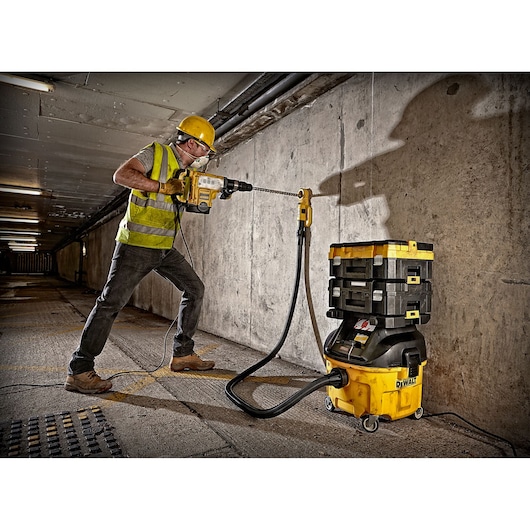 A construction worker wearing a yellow hard hat and reflective vest uses a DEWALT power drill connected to a DEWALT DWV901LT