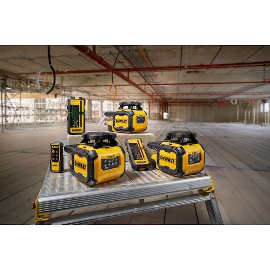 Several DEWALT laser level and measuring tools displayed on a workbench in a construction site. The products are yellow and black and include rotary laser levels and receivers. The background shows an unfinished building with exposed ceilings and scaffolding.