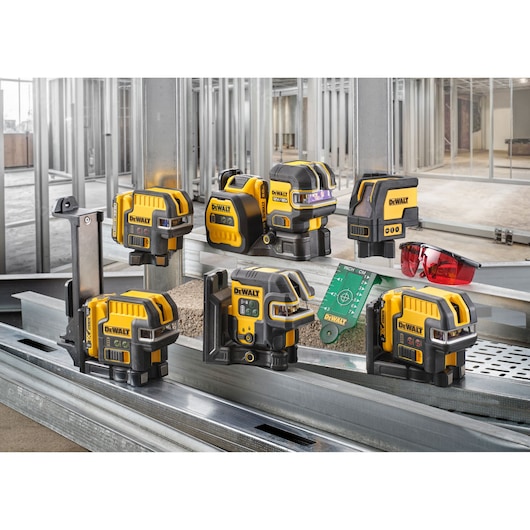 A group of DEWALT yellow and black laser level tools are displayed on metal construction framing, along with a pair of red safety glasses and a green measuring card. The background shows an unfinished building interior with exposed metal studs.