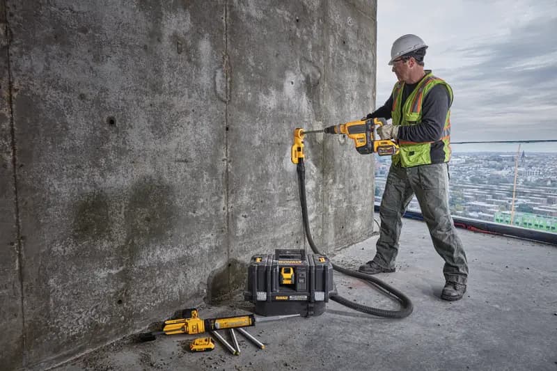 A construction worker wearing a safety vest and helmet is using a DEWALT cordless drill with dust extraction attachment to drill into a concrete wall. The worker is standing on a large building with a cityscape in the background. Several DEWALT tools and a tool case are placed on the ground nearby.