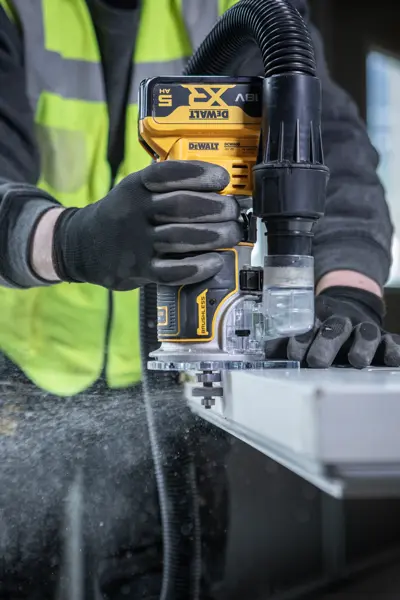 DEWALT DCW604NT-XJ, DCW604NT router in use, worker guides tool on edge with dust extraction. Photography category.