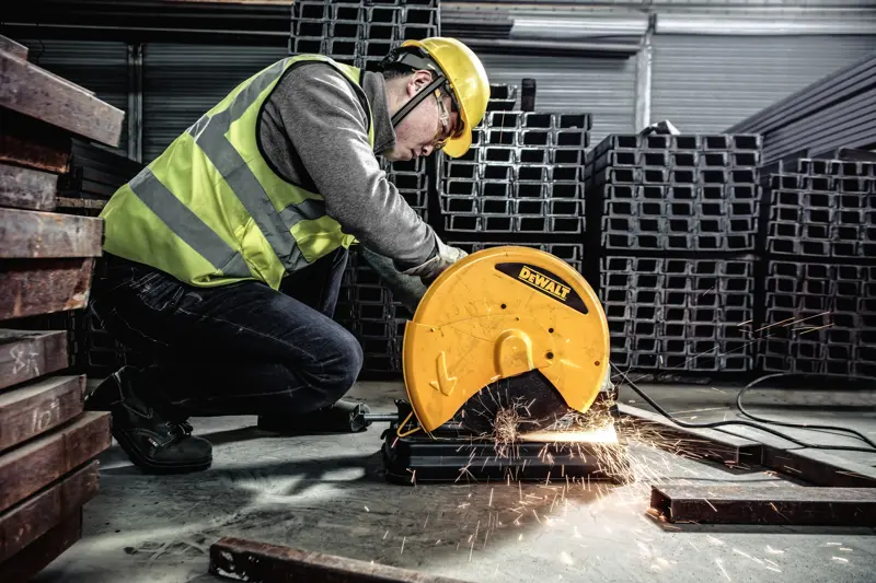 A person wearing safety gear operates a DEWALT metal cutting saw, model D28730, to cut metal bars in an industrial setting. Sparks fly from the cutting area.