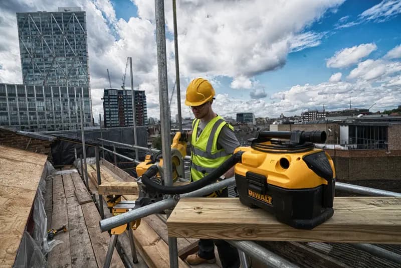 DEWALT DCV584 vacuum on a rooftop construction site with a worker in safety gear and city buildings in the background.