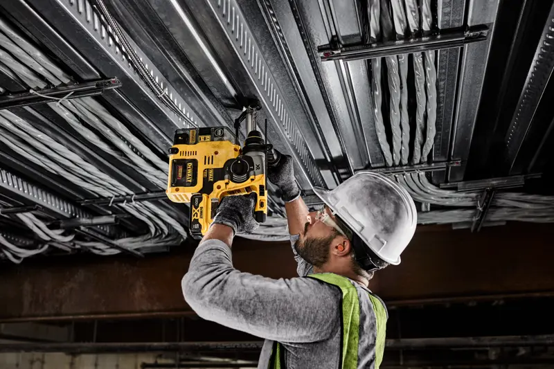 A person wearing a hard hat, safety glasses, gloves, and a safety vest uses a yellow DEWALT cordless rotary hammer drill to drill into a metal ceiling with electrical wiring.