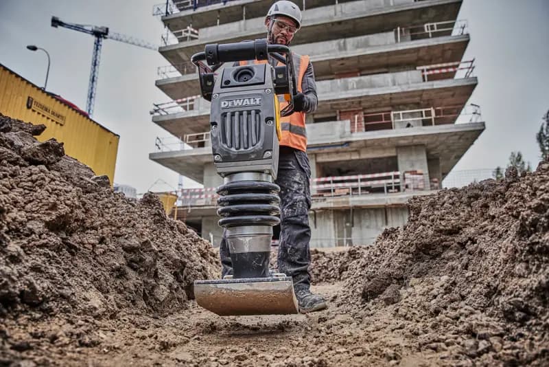DEWALT compactor in use at a construction site, operated by a worker in safety gear, surrounded by soil and buildings.