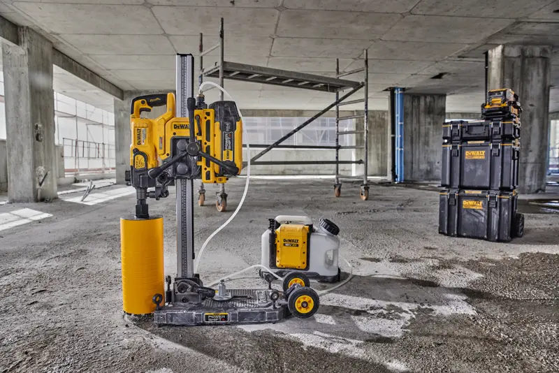 A DEWALT core drilling machine with a yellow cylindrical drill attachment and connected water tank is positioned on the floor of a concrete construction site. In the background, scaffolding and DEWALT branded stackable storage boxes are visible.