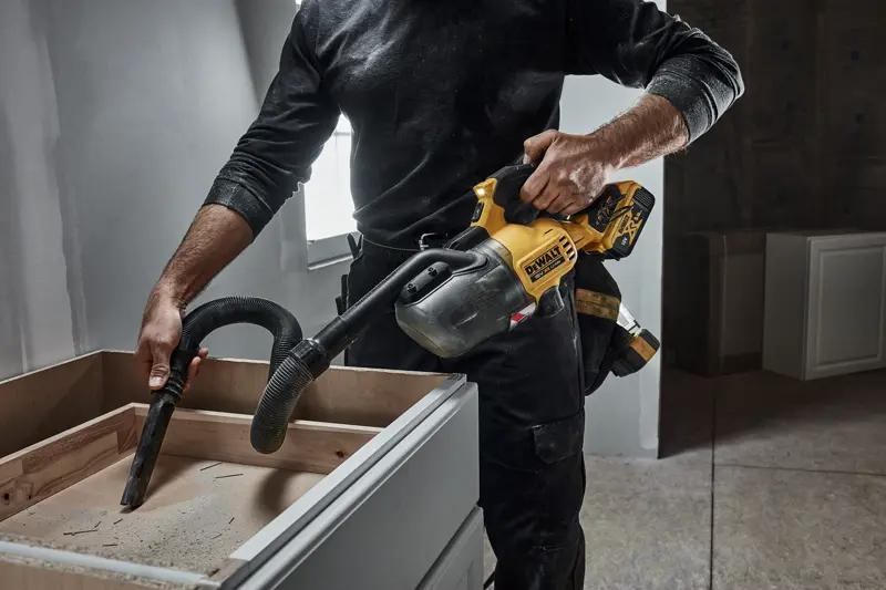 Person uses a DEWALT cordless handheld vacuum to clean dust from a wooden cabinet during construction.