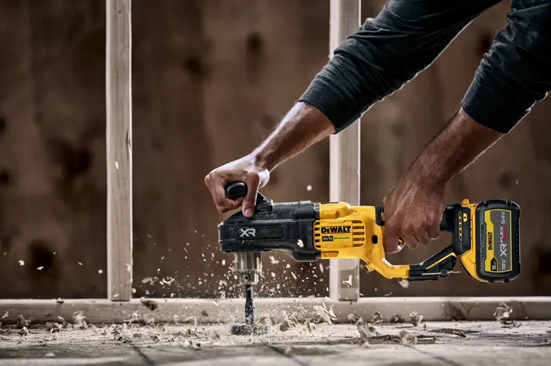 A person using a DEWALT DCD444N cordless power drill with a side handle to drill through wood, creating wood shavings and dust. Construction framing is visible in the background.