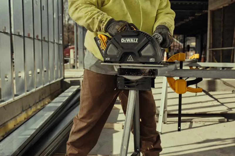 A person wearing work gloves and a yellow sweatshirt is using a DEWALT cordless circular saw to cut metal at a construction site. Sparks are visible as the saw slices through the metal, and the person is operating the saw on a workbench outdoors.
